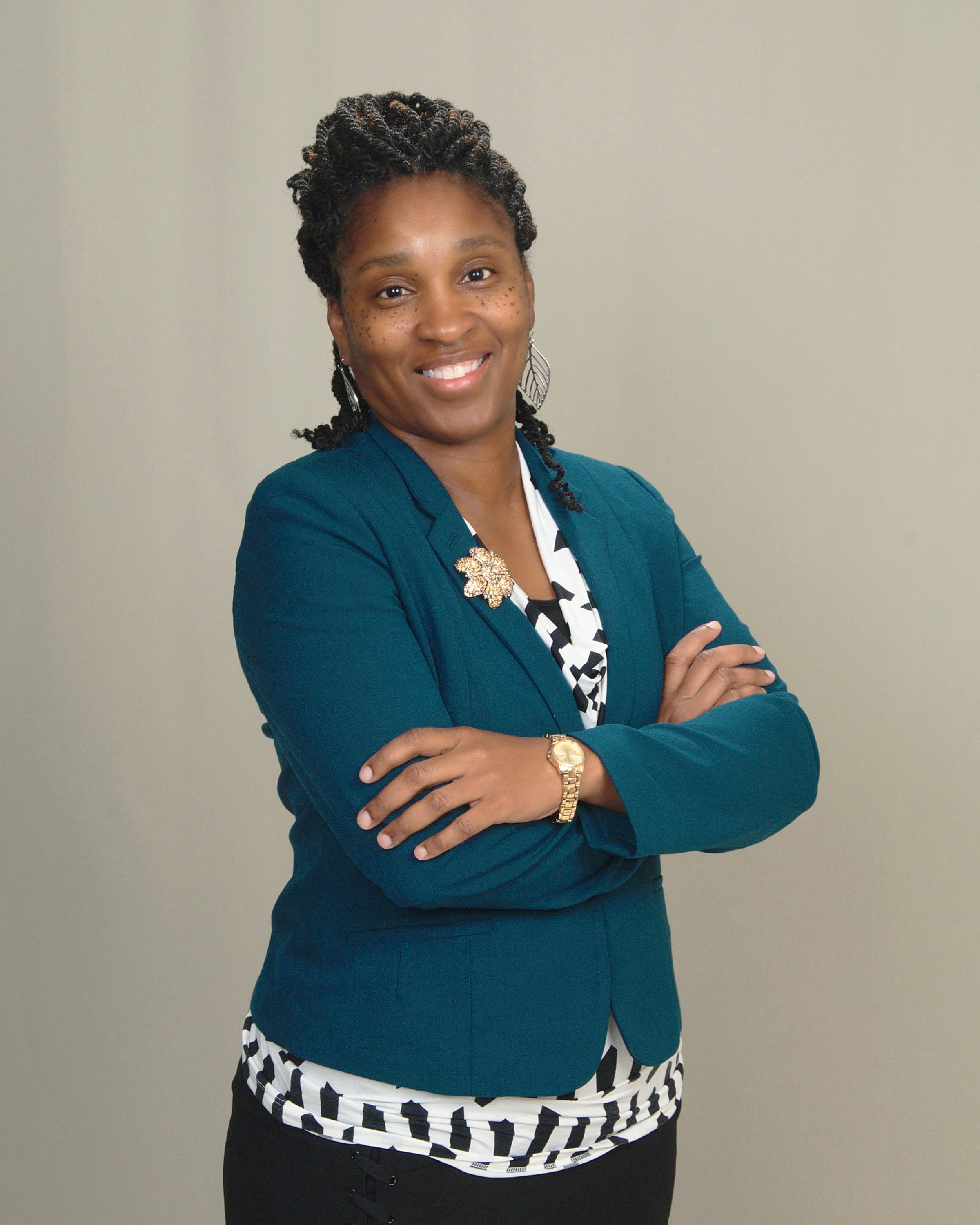 Smiling Black woman with braided hair, teal blazer, and gold jewelry, posing with arms crossed.