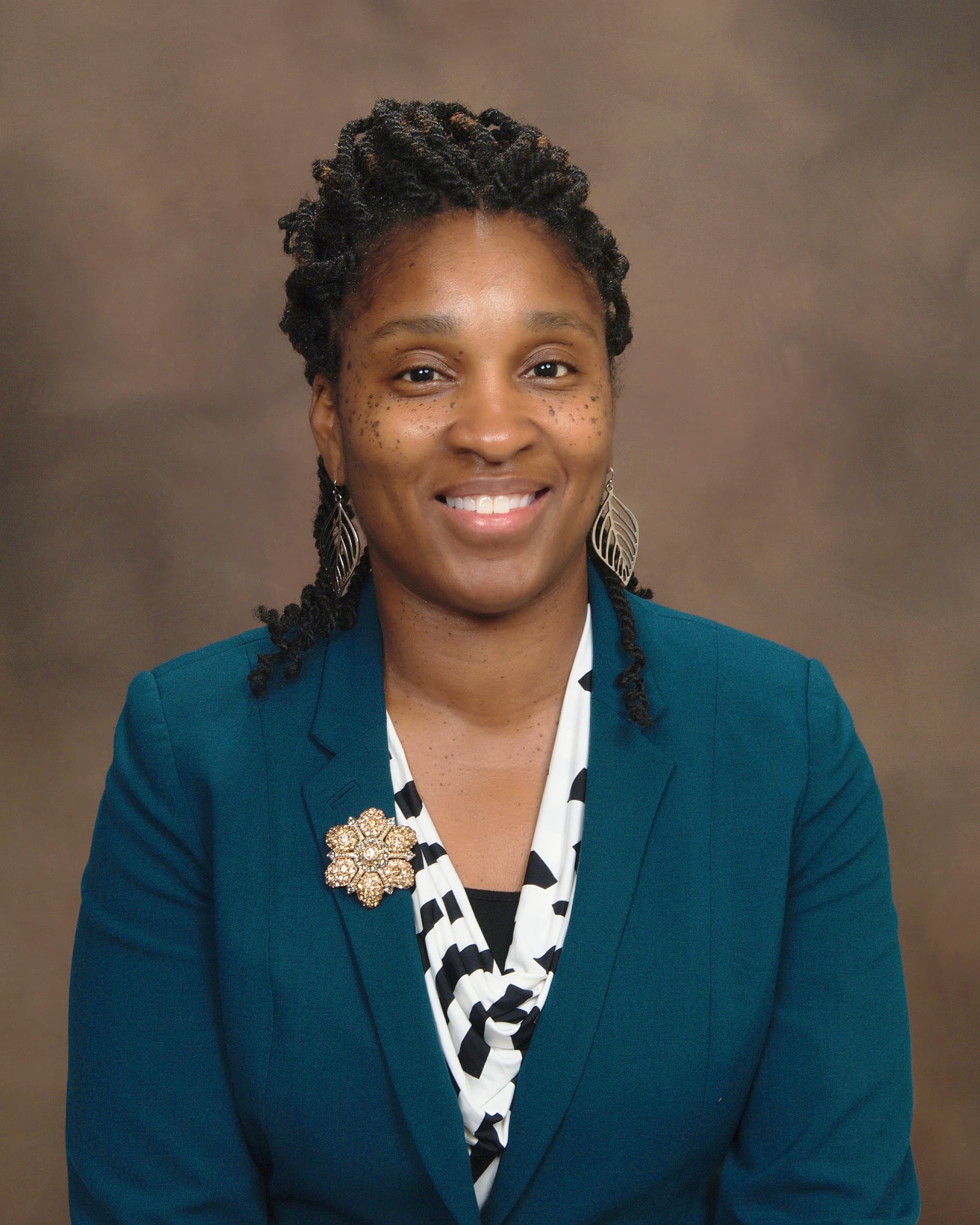 Smiling Black woman with twisted hair wearing a teal blazer and gold flower brooch.